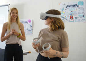 Das Bild zeigt zwei Frauen. Eine Frau hat eine VR-Brille auf und schaut zur Seite. Sie hat zwei Controller in der Hand. Dahinter steht eine Frau und schaut sie an.